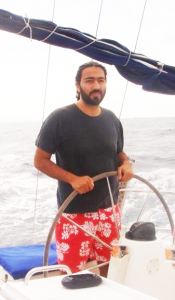 Polivios stands with both hands on the wheel of his sailboat on an overcast day, looking at the camera. He has a dark beard and his hair is pulled back in a ponytail. Behind him, a sail is reefed to the boom and a gently rolling sea stretches back to the horizon. He's wearing a dark t-shirt and red shorts with a white floral pattern.