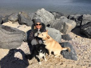 Doreen on a rocky beach with her two dogs. She's wearing winter clothes, sunglasses, and a big grin.