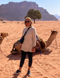 Doreen, a white woman with shoulder-length blonde hair, stands in front of a camel lying down in the sand, with a rock formation and a lone tree in the background. Tire tracks criss-cross the sand underfoot. Doreen wears a light-colored hoodie, black leggings, and sunglasses.